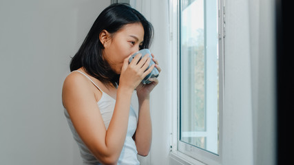 Happy beautiful Asian woman smiling and drinking a cup of coffee or tea near the window in bedroom. Young latin girl open curtains and relax in morning. Lifestyle lady at home concept.