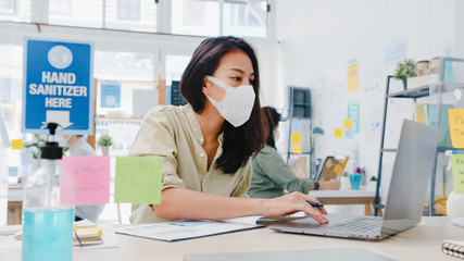Obraz na płótnie Canvas Asia businesswoman entrepreneur wearing medical face mask for social distancing in new normal situation for virus prevention while using laptop back at work in office. Life and work after coronavirus.