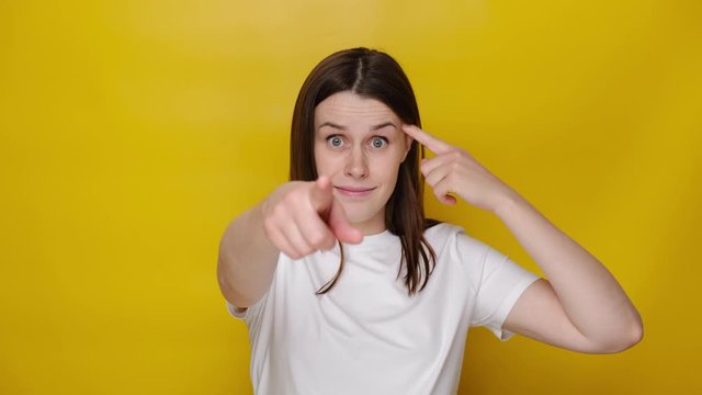 Shocked young woman showing stupid gesture and pointing to camera, accusing crazy cuckoo mind, blaming insane idea, reckless plan, dressed in white t-shirt, isolated on yellow studio background