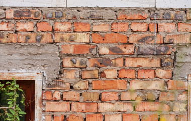 The wall is made of old  red bricks of different shades and cement blocks with a fragment of a basement window. 