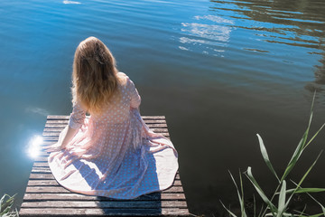 Fototapeta premium Woman on lake. Young woman wearing white dress sitting on a wooden pier on the lake, back view