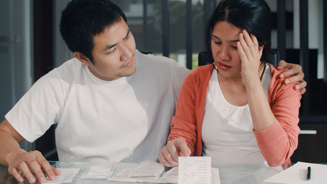 Young Asian Pregnant couple records of income and expenses at home. Mom worried, serious, stress while record budget, tax, financial document working in living room at home concept. - Powered by Adobe