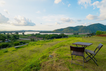 View photos of brown chairs and tables laid on the green lawns on the high mountains in the evening. There is the Mekong River in front and a village in a rural area. Feeling relaxed and calm.
