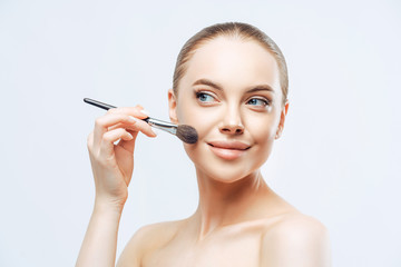 Fototapeta premium Facial treatment, cosmetology concept. Pretty young woman with dark combed hair, applies powder foundation on face with beauty brush, stands sideways, has naked body, isolated on white background