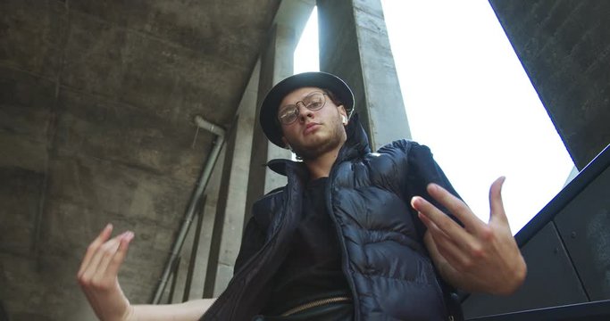 Bottom view of handsome Caucasian boy in glasses and wireless earphones dancing in city. Close up of young funny street dancer in hat doing modern choreography on stairs outdoor. Hip hop dance concept