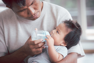 Obraz na płótnie Canvas Asian father holding and feeding her baby boy with milk from baby bottle.