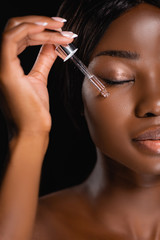 portrait of african american naked woman applying serum on face isolated on black