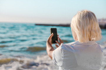 Portrait caucasian blond woman in striped shirt makes selfie photo by smartphone of the sea on a sunny day.