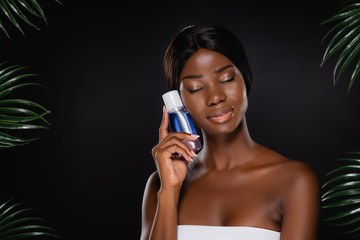 Fototapeta premium african american woman with closed eyes holding micellar water near green palm leaves isolated on black