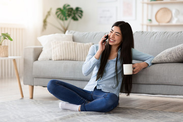 Free Time Leisure. Joyful Muslim Girl In Headscarf Talking On Cellphone With Friend While Relaxing At Home With Coffee, Copy Space