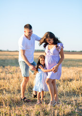 happy young family on the field, pregnant wife and a little girl hugs with father, wind turbines on the background