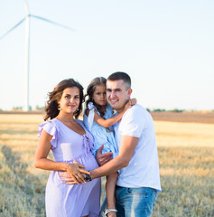 happy young family on the field, pregnant wife and a little girl hugs with father, wind turbines on the background