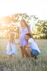 Naklejka na ściany i meble happy young family on the field in the sunset light, husband and a little girl are kissing pregnant wife belly