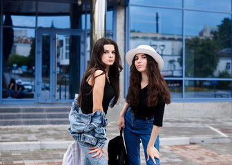 Two young brunette girls, wearing casual jeans attire, standing in front of blue modern glass building. Girlfriends traveling in city, holding backpacks, sightseeing. Active lifestyle concept.