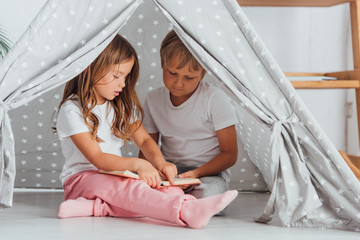 brother and sister in pajamas sitting on floor in kids wigwam and reading book