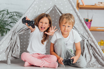 KYIV, UKRAINE - JULY 21, 2020: excited girl with outstretched hands looking at camera while playing video game with brother near kids wigwam © LIGHTFIELD STUDIOS