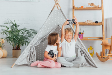 KYIV, UKRAINE - JULY 21, 2020: boy showing winner gesture near sister holding joystick while sitting on floor near kids wigwam © LIGHTFIELD STUDIOS