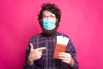 Photo of curly man wearing medicinal mask and pointing at passport with avia tickets.