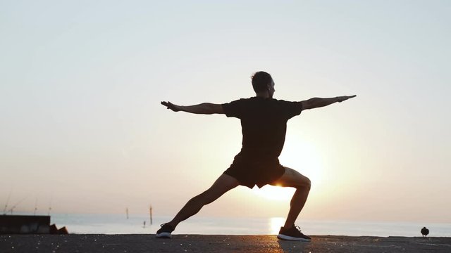 Morning workout. Warm up and stretching before training on the sea pier. Young man practicing youga against the backdrop of the sea and the rising sun. Sport activity. Athlete silhouette. 