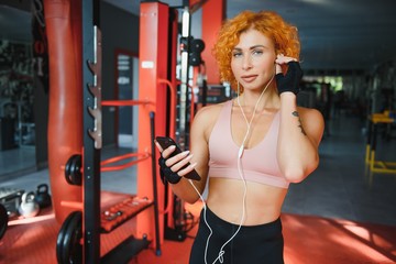 Smiling charming young woman athlete in earphones using smartphone and listening to music in gym