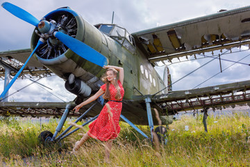 Young nice woman in a red pin up style dress stands by an abandoned green plane