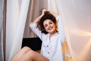 Fototapeta premium A cheerful, attractive girl sits on a chair, an armchair with a smile in a white shirt, sexy, cute, holds her hair
