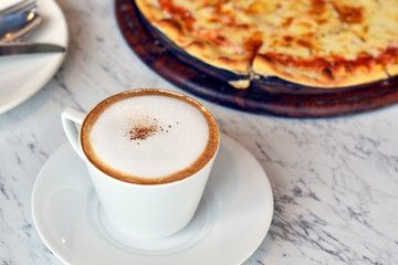 Hot coffee in white cup on marble table with blurred pizza background.