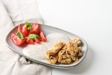 Pieces of oriental marinated chicken, yoghurt sesame sauce and tomatoes with basil garnish, served on a small plate on a white table, copy space