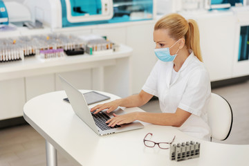 Beautiful blond female lab assistant sitting in lab with protective mask on and using laptop during covid outbreak.