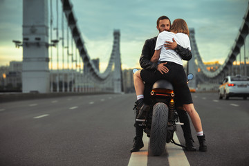 Young sexy woman hugging cute man in stylish black leather jacket, sitting on sports motorcycle on the bridge in the city on sunset and kissing