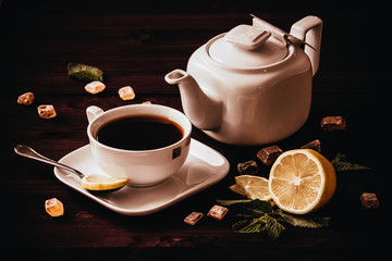 
A cup of tea with lemon and a teapot. Lemon and cubes of natural sugar lie on a dark wooden table