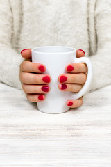 Female hands hold a white cup against the background of a white fluffy sweater with a beautiful red winter manicure. Drink, fashion, morning. New Year. Celebration. Tea coffee.