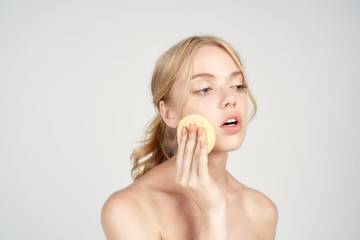 A blonde with bare shoulders holds cotton pads near her face skin care light background