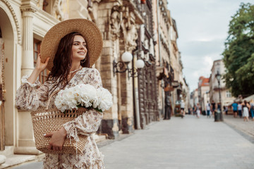 Fototapeta na wymiar Happy smiling lady wearing straw hat, floral print dress, holding wicker bag with white peony flowers, posing in street of European city. Lifestyle, travel conception. Copy, empty space for text 