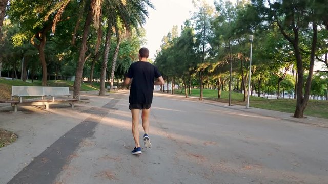 Morning workout. Young man is running in the sunny urban park. Sport activity. Athlete. Jogging in the city. Man in black t-shirt training among trees and palms. Healthy lifestyle.