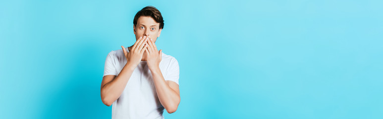 Panoramic orientation of shocked man covering mouth with hands on blue background