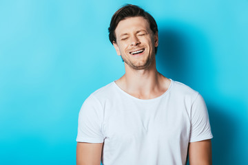 Young man in white t-shirt laughing with closed eyes on blue background