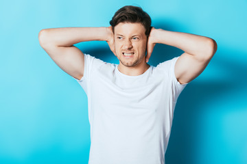 Fototapeta na wymiar Angry man in white t-shirt covering ears with hands on blue background
