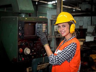 Portrait of confident young female mechanical engineer in a protective uniform, helmet, and sound protector controls the CNC Machinery while working at the plant.