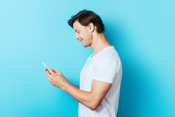 Side view of young man in white t-shirt using smartphone on blue background