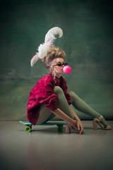 Blowing bubble with gum on skateboard. Young woman as Marie Antoinette on dark green background....