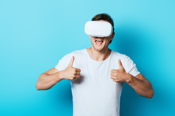 Naklejka premium Young man showing thumbs up while using virtual reality headset on blue background