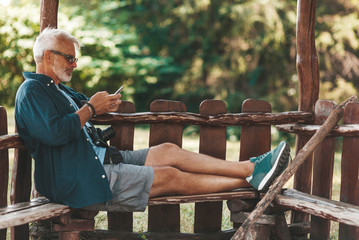An elderly man is typing a message on the phone while relaxing on a bench. Active lifestyle in old age. Spending time in the fresh air.
