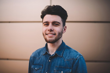 Half length portrait of smiling young man with trendy hairstyle dressed in casual blue denim shirt, close up of male's face, positive bearded hipster guy looking at camera standing on wall background