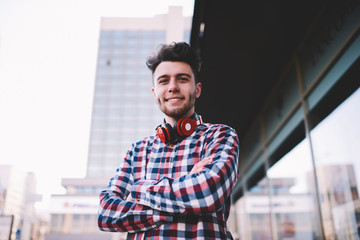 Half length portrait of confident hipster guy standing with crossed arms outdoors, handsome positive young man in stylish shirt and with modern red headphones looking at camera posting