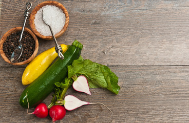 top view, close distance of freshly picked vegetables of yellow, green squash, halved and whole reddish with leaves and a rare wood bowel of sea salt with an artisan spoon on a wood table top