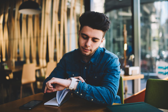 Pensive Young Man Checking Notification On Modern Wearable Smartwatch Reading Message On Touch Screen, Hipster Guy Checking Time On Modern Digital Watch With Organizer  Sitting In Cafe Interior