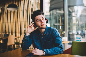 Dreamy young man enjoying playlist via cellular and headphones connected with bluetooth, handsome hipster guy sitting at cafe interior listening to audio book via electronic accessories and telephone