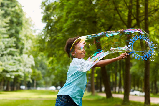 Waist Up Portrait Of Cute African-American Boy Playing With Bubbles Outdoors In Green Park Holding Big Bubble Wand, Copy Space