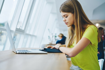 Pensive beautiful student dressed in yellow t-shirt reading records written in notepad sitting in cafeteria indoors with modern laptop computer connecting to wireless internet.Advertising area